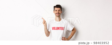 Handsome smiling volunteer in t-shirt pointing finger left, showing announcement, standing over white background 98403952