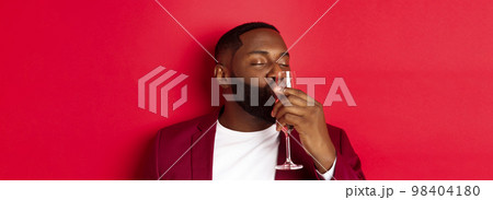 Drunk african american guy kissing glass with champagne, celebrating new year, enjoying party, standing over red background 98404180