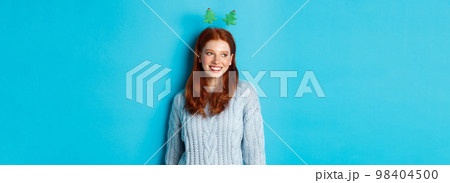 Winter holidays and Christmas sales concept. Cute redhead girl in funny New Year headband smiling, looking left at logo, standing over blue background 98404500