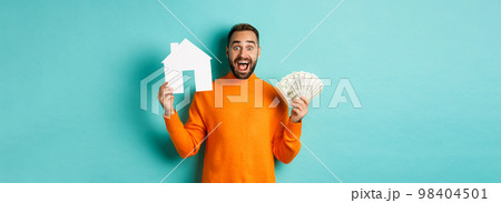 Real estate concept and mortgage concept. Excited man showing dollars and paper maket of house, buying flat, standing over blue background Real estate concept and mortgage concept. Excited man showing dollars and paper maket of house, buying flat, standing over blue background 98404501