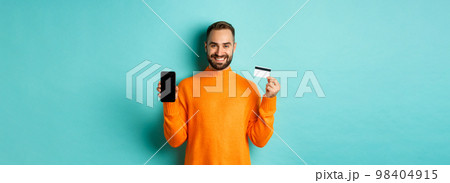 Online shopping. Happy attractive guy showing mobile phone screen and credit card, smiling satisfied, standing over light blue background 98404915