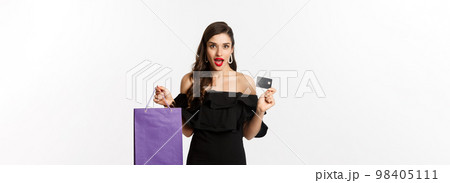 Stylish young woman in black dress going shopping, holding bag and credit card, smiling pleased, standing over white background 98405111
