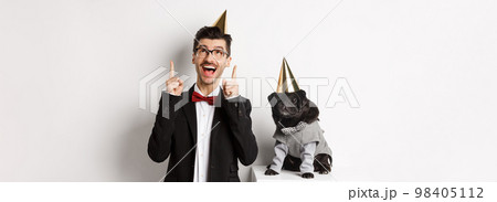 Cute black pug dog wearing party cone and standing near happy owner, man pointing fingers up at copy space, celebrating birthday, white background 98405112