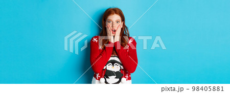 Winter holidays and Christmas Eve concept. Impressed and speechless redhead girl gasping, staring at camera with disbelief, standing in xmas sweater against blue background Winter holidays and Christmas Eve concept. Impressed and speechless redhead girl gasping, staring at camera with disbelief, standing in xmas sweater against blue background 98405881