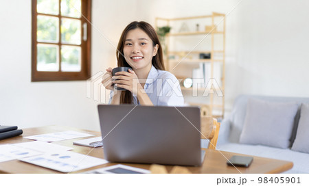 Beautiful Asian woman working on laptop and sipping coffee with smiling face in her home, Creating happiness at work with a smile, Freelancer working at home happily, Work from home. 98405901