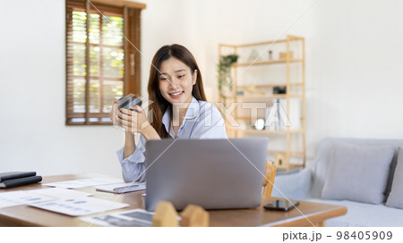 Beautiful Asian woman working on laptop and sipping coffee with smiling face in her home, Creating happiness at work with a smile, Freelancer working at home happily, Work from home. Beautiful Asian woman working on laptop and sipping coffee with smiling face in her home, Creating happiness at work with a smile, Freelancer working at home happily, Work from home. 98405909