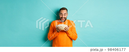 Handsome bearded man looking with exctied and happy at popcorn bowl, ready to watch movie, standing over white background Handsome bearded man looking with exctied and happy at popcorn bowl, ready to watch movie, standing over white background 98406177