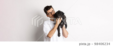 Handsome young man pet his cute black dog, scratching pug while holding animal on shoulder, standing over white background 98406234