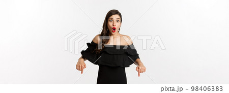 Fashion and beauty. Elegant woman in black dress pointing fingers down, showing promo and smiling, standing over white background 98406383