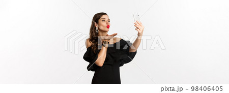 Stylish young woman in black dress, partying and taking selfie on mobile phone, sending air kiss at smartphone camera, standing over white background 98406405