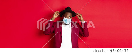 Covid-19, quarantine and holidays concept. Handsome and stylish african american man in face mask, put hat on head and looking sassy, dress-up for party, standing over red background Covid-19, quarantine and holidays concept. Handsome and stylish african american man in face mask, put hat on head and looking sassy, dress-up for party, standing over red background 98406435