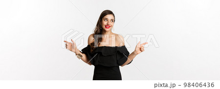Fashion and beauty. Attractive woman in jewelry, makeup and black dress, smiling and pointing fingers sideways copy space offer, white background 98406436