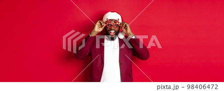 Funny Black man celebrating New Year, wearing party glasses and santa hat, having fun over red background Funny Black man celebrating New Year, wearing party glasses and santa hat, having fun over red background 98406472
