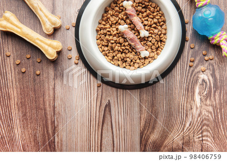 A bowl with dog food, dog treats and toys on a wooden floor. 98406759