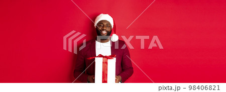 Christmas, New Year and shopping concept. Happy bearded african american man holding xmas present, smiling at camera, standing in santa hat against red background 98406821
