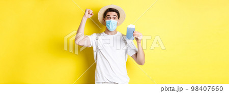 Concept of covid-19, travelling and quarantine. Happy man tourist in medical mask celebrating, showing passport with tickets for vacation and rejoicing, travel during coronavirus 98407660