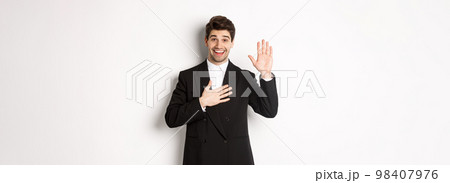 Image of handsome smiling guy telling the truth, raising on arm to promise something, standing over white background in black suit Image of handsome smiling guy telling the truth, raising on arm to promise something, standing over white background in black suit 98407976