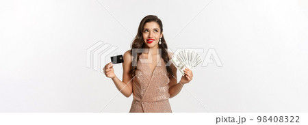 Shopping concept. Young woman deciding what to buy, looking thoughtful at upper left corner and showing credit card with money, standing in party dress Shopping concept. Young woman deciding what to buy, looking thoughtful at upper left corner and showing credit card with money, standing in party dress 98408322