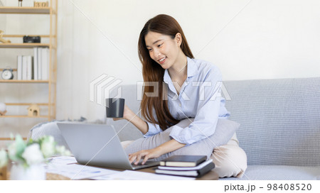 Beautiful Asian woman working on laptop and sipping coffee with smiling face in her home, Creating happiness at work with a smile, Freelancer working at home happily, Work from home. 98408520