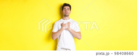 Romantic and nostalgic man holding hands on heart, close eyes and remember something, standing over yellow background 98408660