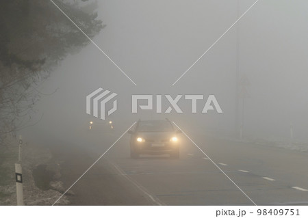 Car traveling on the foggy road with headlights or headlamps on. Low visibility - Dangerous driving of cars in winter in bad weather. 98409751