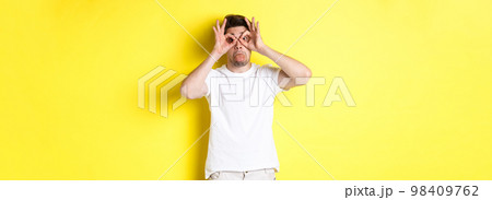 Young man making funny faces and showing tongue, fool around, standing in white t-shirt against yellow background 98409762