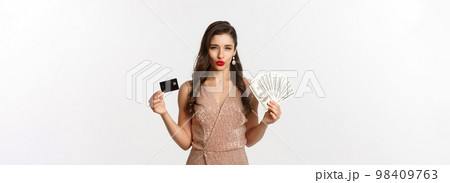 Shopping concept. Sassy woman in elegant dress showing credit card with money, looking pleased at camera, standing over white background Shopping concept. Sassy woman in elegant dress showing credit card with money, looking pleased at camera, standing over white background 98409763
