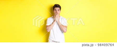Handsome man looking shocked and speechless, holding hands on mouth, standing over yellow background 98409764