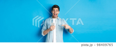 Concept of online shopping, applications and technology. Happy smiling man pointing finger at phone, standing amazed against blue background Concept of online shopping, applications and technology. Happy smiling man pointing finger at phone, standing amazed against blue background 98409765
