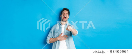 Adult man in casual outfit laughing out loud and pointing at you, looking at something funny, chuckling while standing against blue background Adult man in casual outfit laughing out loud and pointing at you, looking at something funny, chuckling while standing against blue background 98409964