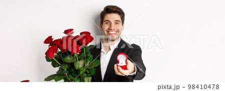 Close-up of attractive man in suit, holding bouquet of roses giving an engagement ring, proposing to girlfriend, standing against white background Close-up of attractive man in suit, holding bouquet of roses giving an engagement ring, proposing to girlfriend, standing against white background 98410478