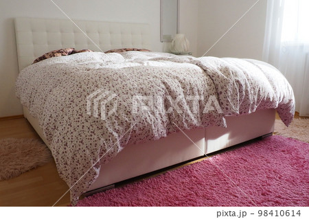 Cozy compact bedroom with white furniture, pink carpet and tulle curtains. Fluffy feather blanket is thrown over the bed. Romantic morning. Double bed with upholstered headboard. Light from the window 98410614