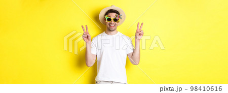 Concept of tourism and lifestyle. Happy man enjoying trip, wearing summer hat and sunglasses, posing with peace signs for photo, yellow background Concept of tourism and lifestyle. Happy man enjoying trip, wearing summer hat and sunglasses, posing with peace signs for photo, yellow background 98410616