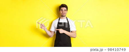 Doubtful barista in black apron pointing fingers right, looking skeptical and unamused, standing over yellow background 98410749