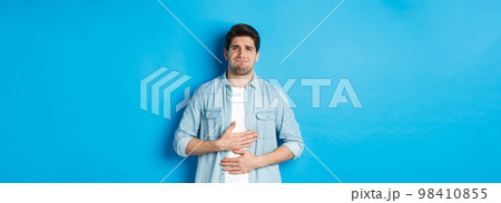 Man holding hands on belly and grimacing from pain, complaining on stomach ache, standing against blue background Man holding hands on belly and grimacing from pain, complaining on stomach ache, standing against blue background 98410855