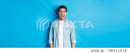 Attractive happy man in glasses looking surprised, checking out advertisement, standing over blue background 98411018