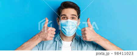 Concept of covid-19, pandemic and quarantine. Close-up of cheerful young man in medical mask smiling, showing thumbs up in approval, like and agree, standing over blue background 98411023