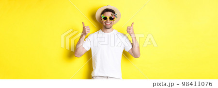 Concept of tourism and lifestyle. Image of smiling tourist showing thumbs-up, enjoying trip and recommending, wearing summer hat and sunglasses, yellow background 98411076