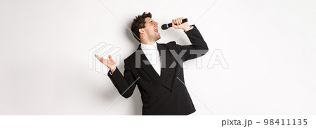 Portrait of handsome man singing a song with passion, standing in black suit, holding microphone and performning, posing over white background Portrait of handsome man singing a song with passion, standing in black suit, holding microphone and performning, posing over white background 98411135