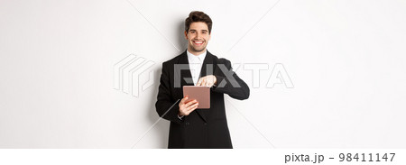 Portrait of handsome, stylish male entrepreneur in black suit pointing at digital tablet, showing something online, standing against white background 98411147