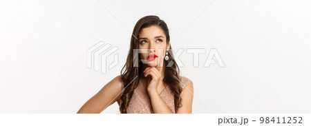 Concept of New Year celebration and winter holidays. Close-up of elegant woman with red lips and dress, looking at upper left corner and thinking, standing over white background 98411252