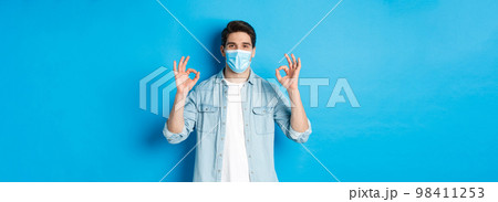 Concept of covid-19, pandemic and social distancing. Satisfied man in medical mask, showing ok signs in approval, agree or like something good, standing over blue background 98411253