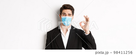 Concept of covid-19, business and social distancing. Close-up of good-looking businessman in trendy suit and medical mask, showing okay sign and winking, standing against white background 98411256