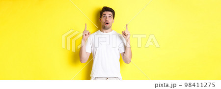 Impressed guy checking out promo offer, looking and pointing fingers up at advertisement, standing over yellow background 98411275