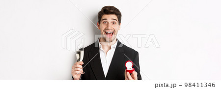 Close-up of handsome man in suit, making a proposal, giving engagement ring and raising glass of champagne, standing against white background Close-up of handsome man in suit, making a proposal, giving engagement ring and raising glass of champagne, standing against white background 98411346