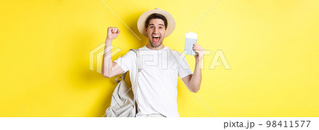 Tourism and vacation. Man feeling happy about summer trip, holding passport with plane tickets and backpack, raising hands up in celebration gesture, yellow background Tourism and vacation. Man feeling happy about summer trip, holding passport with plane tickets and backpack, raising hands up in celebration gesture, yellow background 98411577