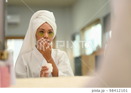 Gorgeous woman in bathrobe using under eye patches, doing morning routine after shower. Self care and daily beauty routine 98412101