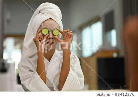 Portrait of millennial woman with facial mask on her face holding slices of fresh cucumber. Reflecting in mirror of toilette dresser table 98412276