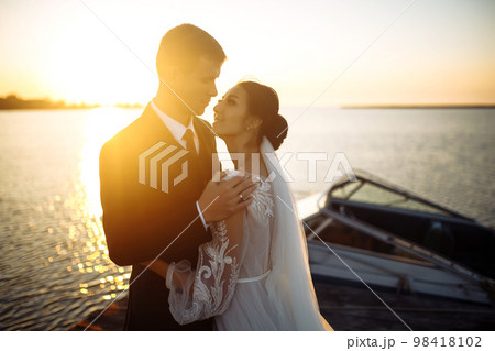 Beautiful bride and stylish groom together on the bridge against the background of the boat at sunset. 98418102