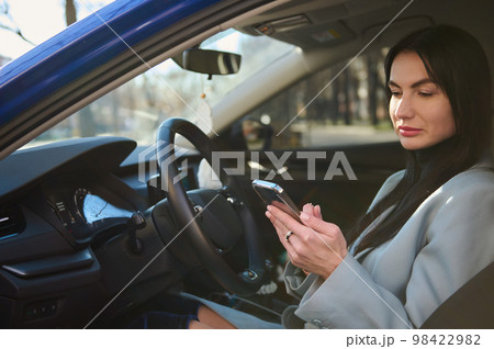 Confident successful middle-aged businesswoman, dressed in winter coat, sitting in the car on driver's seat, and using smart phone, making call to clients, planning business meetings. People Business 98422982